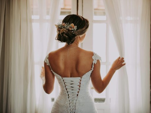 woman in wedding dress standing near window looking outside while holding the curtains