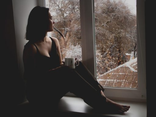 Vrouw zit bij het raam en kijkt peinzend naar buiten, zichtbaar gestrest